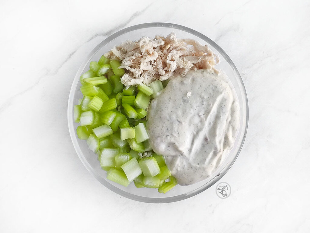 A clear glass bowl with three distinct sections ready to be mixed: shredded chicken, a pile of diced celery, and a large dollop of thick, creamy ranch dressing.
