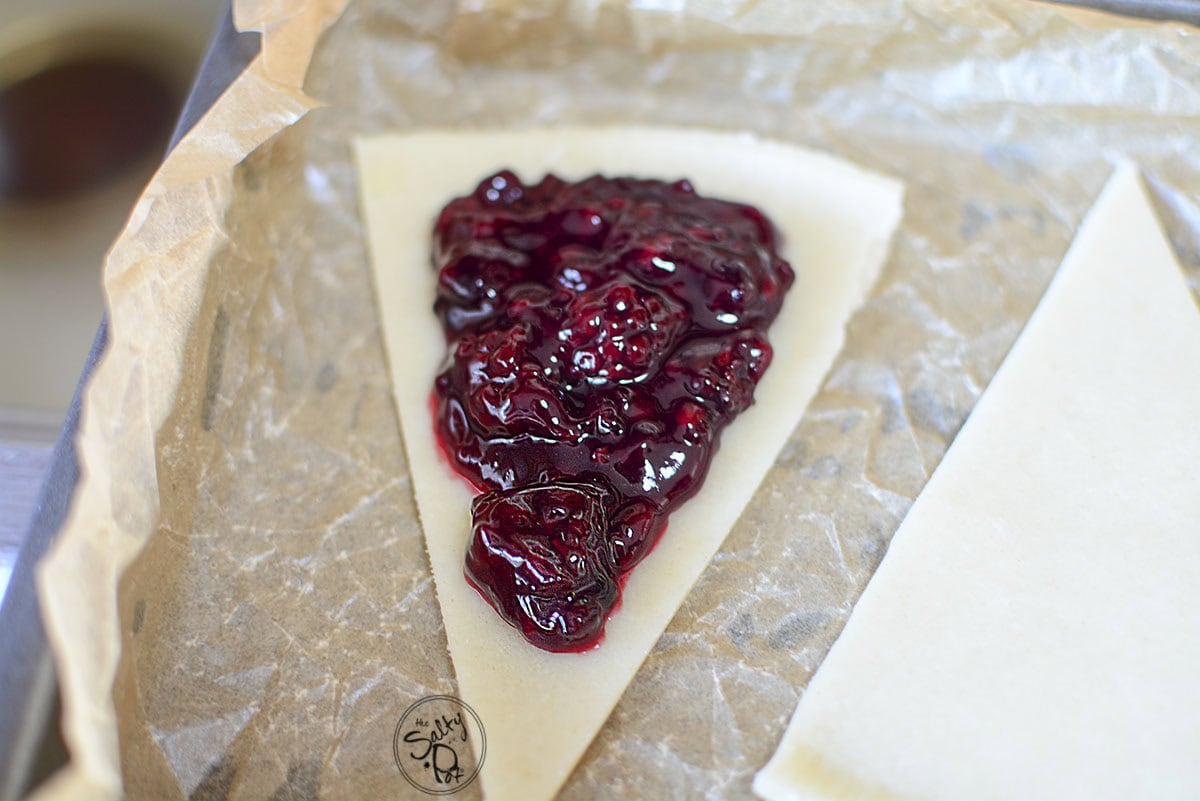 A single triangle of pie dough on parchment paper topped with a generous spoonful of thick, dark purple blackberry filling, leaving space around the edges for sealing.