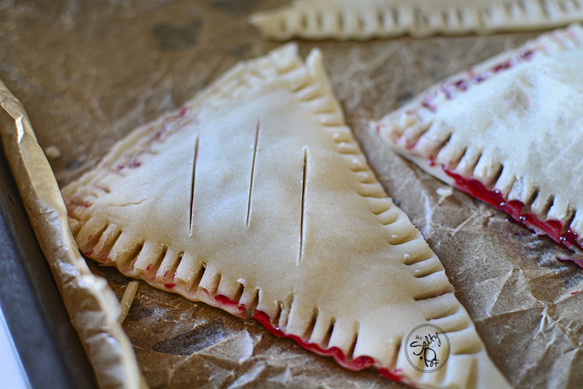A close-up of unbaked triangle-shaped hand pies on a parchment-lined baking sheet. The edges are crimped with a fork, and three small slits are cut into the top of the dough.