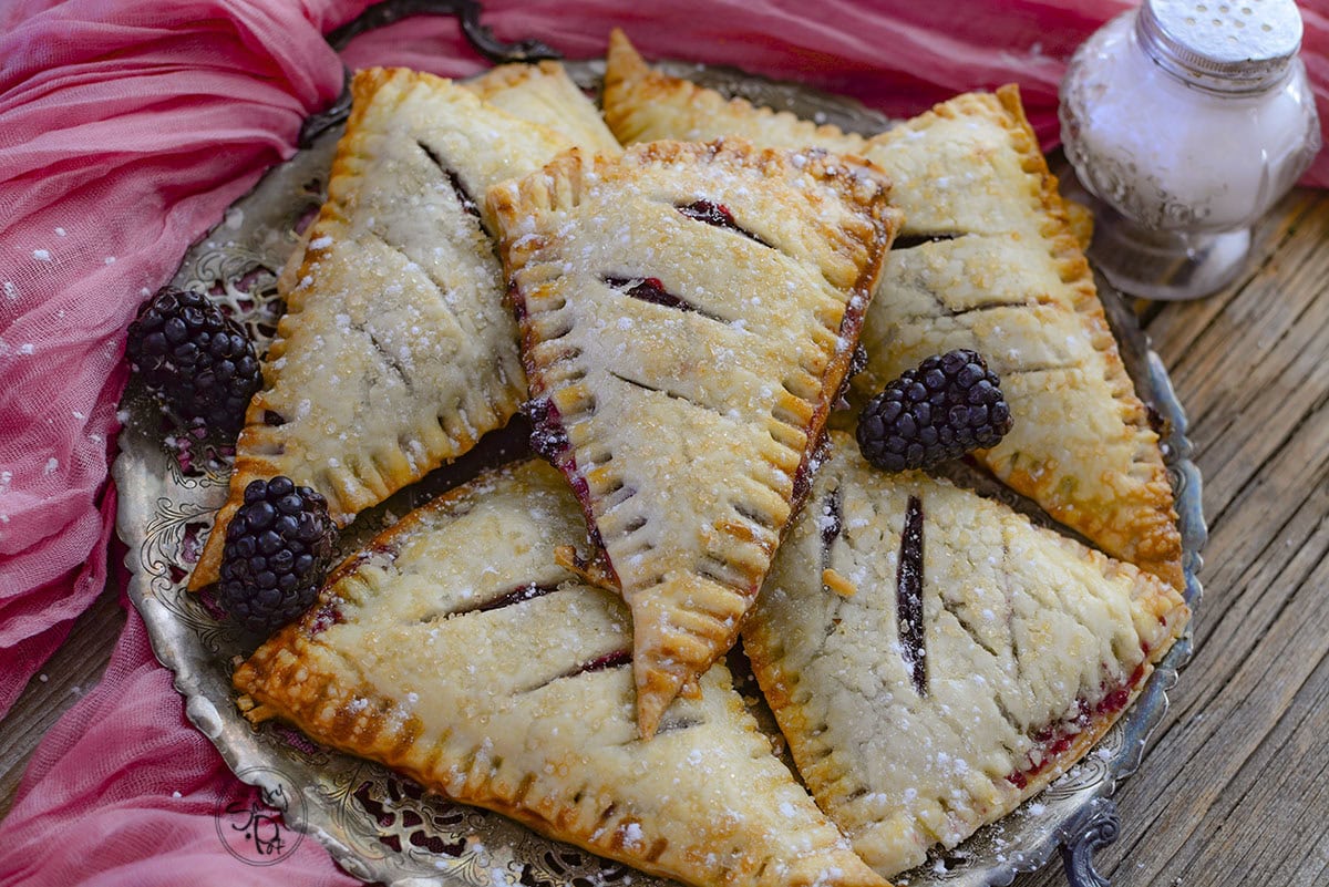 A close-up shot of golden-brown baked blackberry hand pies stacked on a silver platter. They are dusted with powdered sugar and garnished with fresh blackberries against a pink fabric background.