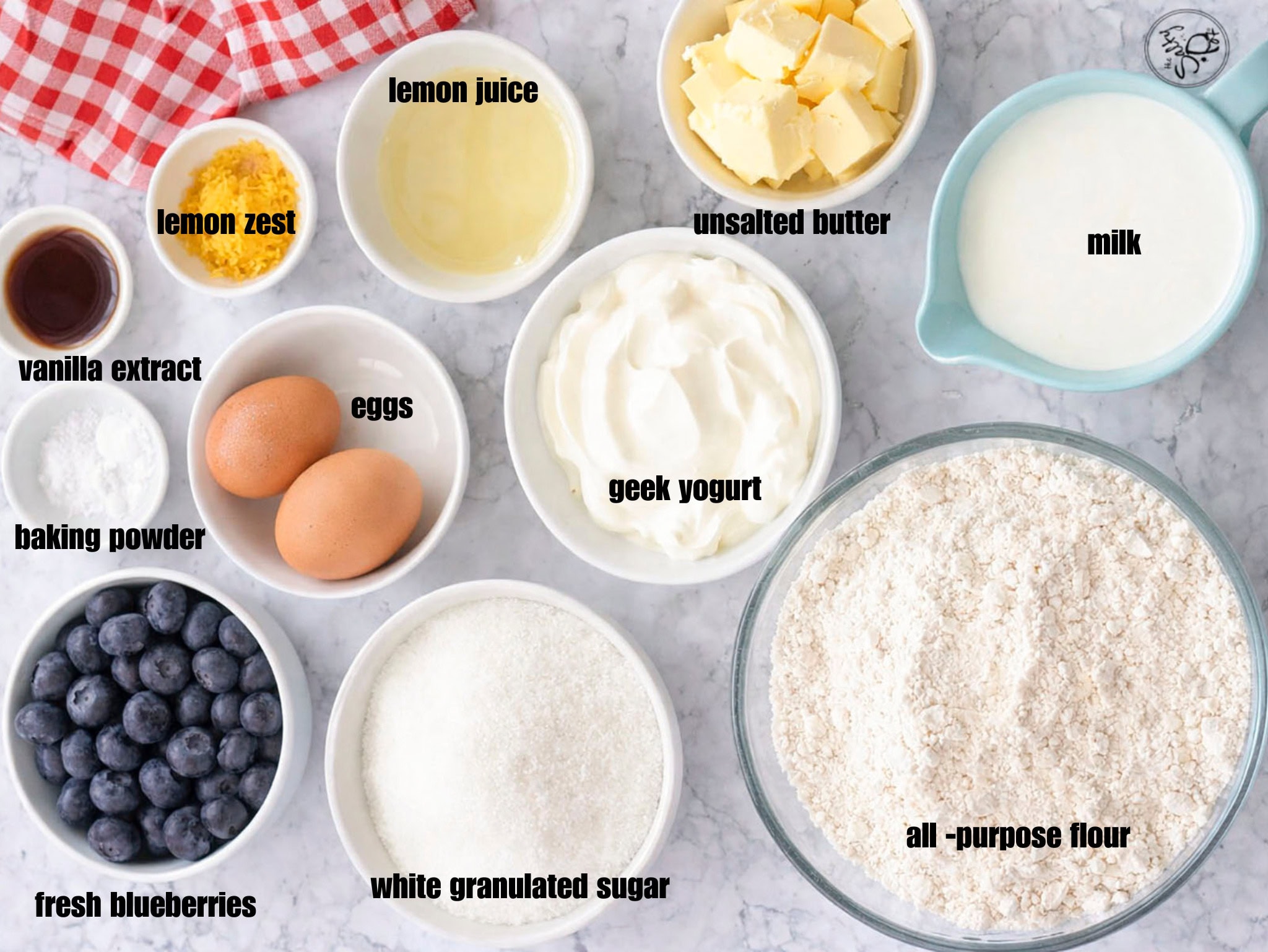 A flat lay of all the cake ingredients in small white bowls, including fresh blueberries, lemon zest, Greek yogurt, and cubes of butter on a marble background.
