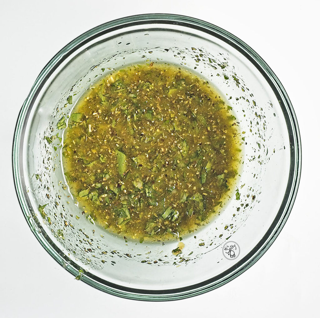 A glass mixing bowl showing the salad dressing after being whisked together. The mixture is a vibrant green and gold emulsified vinaigrette with visible bits of chopped herbs and spices.