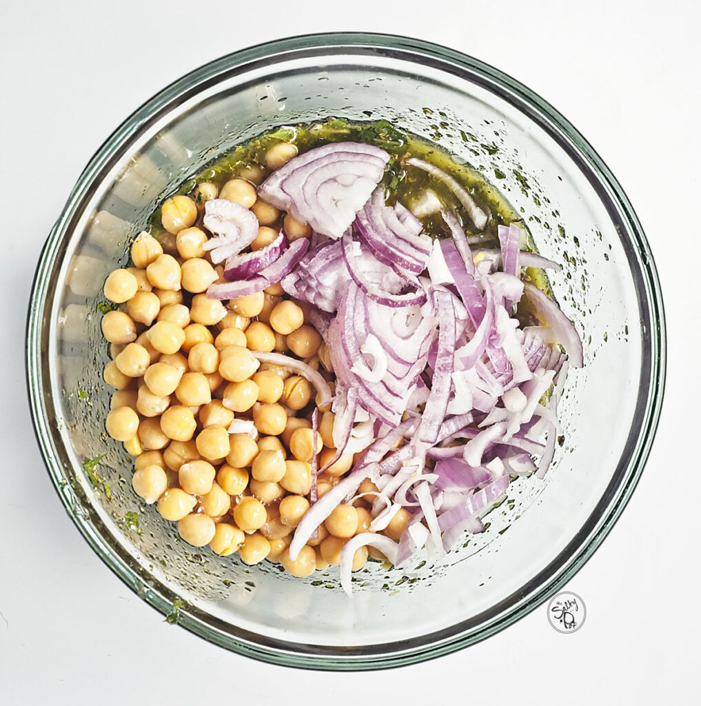 A glass bowl containing the herb vinaigrette with whole chickpeas and thinly sliced red onions added on top, ready to be tossed.