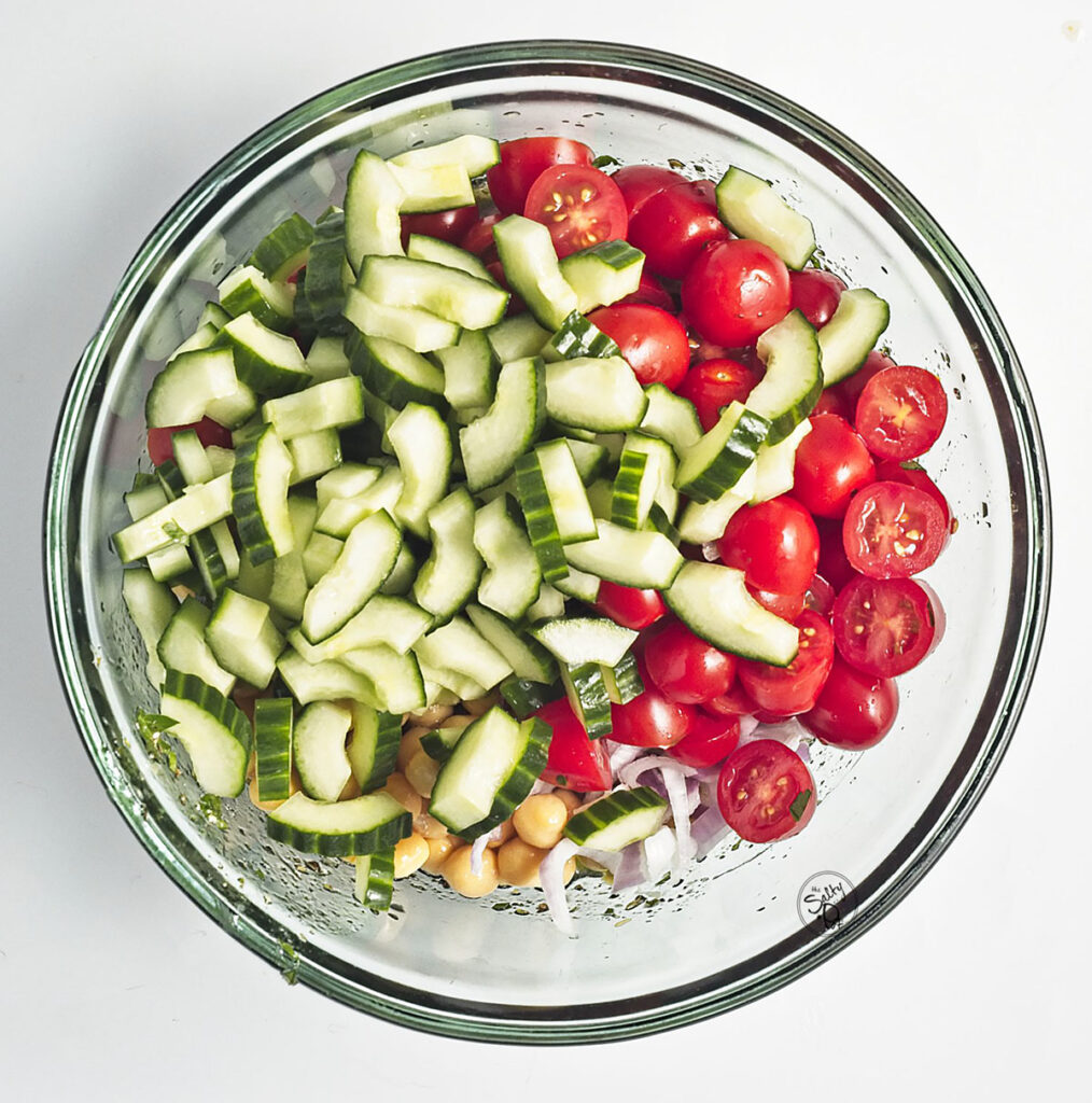 The glass bowl now contains chickpeas, red onions, halved cherry tomatoes, and chopped cucumbers layered over the dressing.