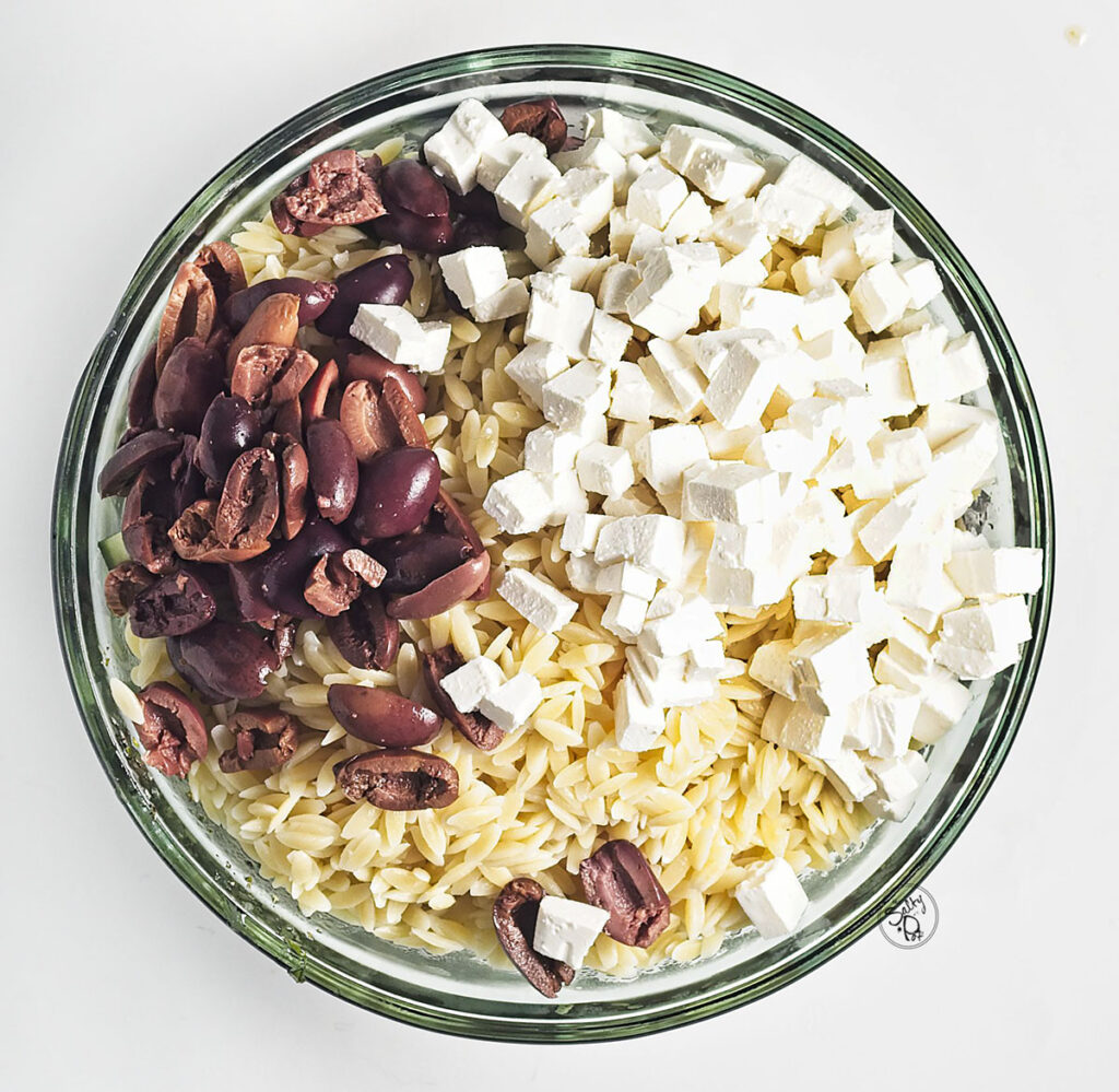 A top-down view of the glass bowl filled with cooked orzo pasta, topped with a large portion of cubed white feta cheese and pitted Kalamata olives.