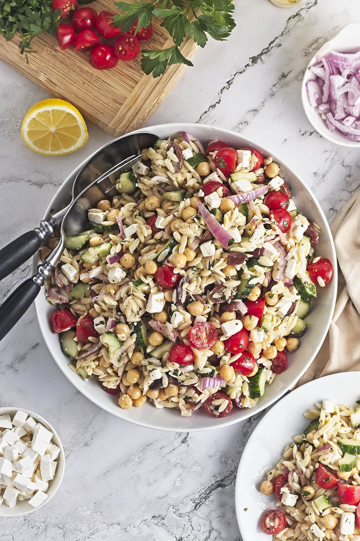 A large white serving bowl filled with the completed Chickpea Orzo Salad. Two large serving spoons rest in the bowl. Surrounding the bowl are garnishes including a lemon half, extra feta cheese, sliced red onions, and fresh tomatoes on a wooden cutting board.