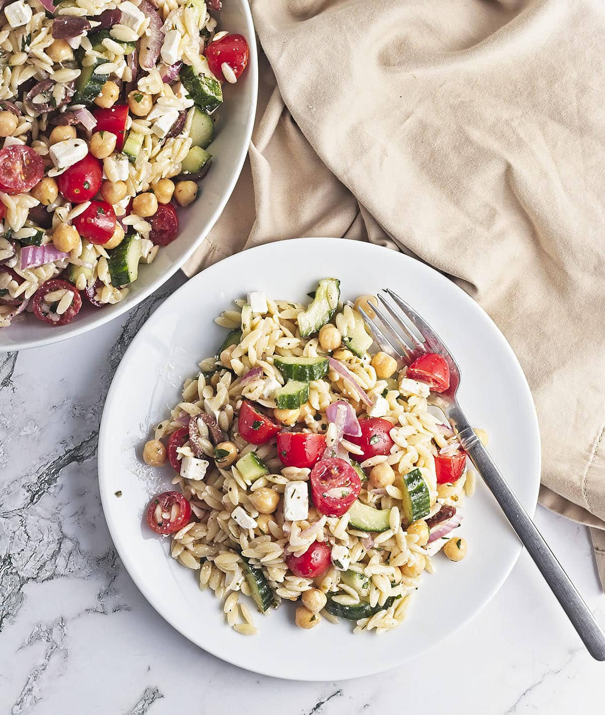 A close-up, top-down shot of a single serving of the Chickpea Orzo Salad on a white plate with a silver fork. The salad clearly shows the textures of the orzo, chickpeas, feta, cucumbers, and tomatoes coated in herb dressing.