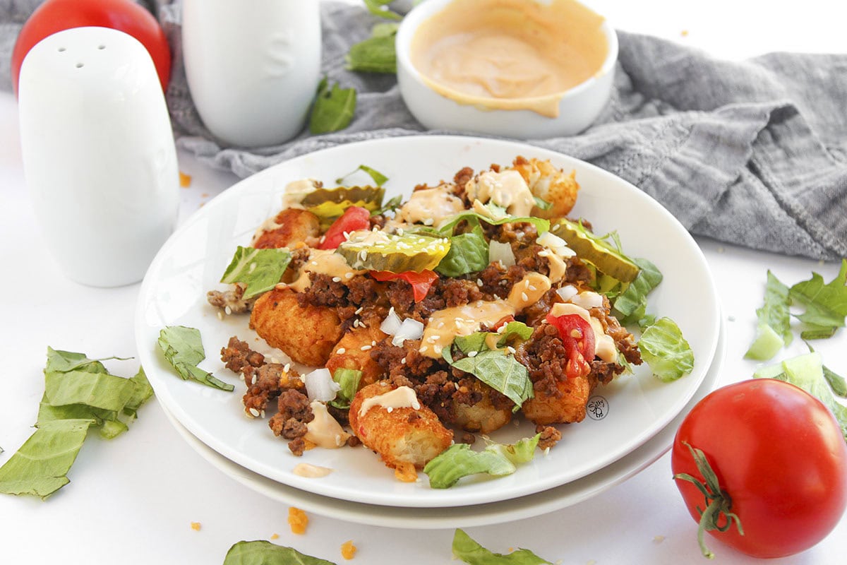 A white plate featuring a generous serving of cheeseburger-style loaded tater tots with melted cheese, beef, fresh toppings, and a side bowl of burger sauce on a marble countertop.