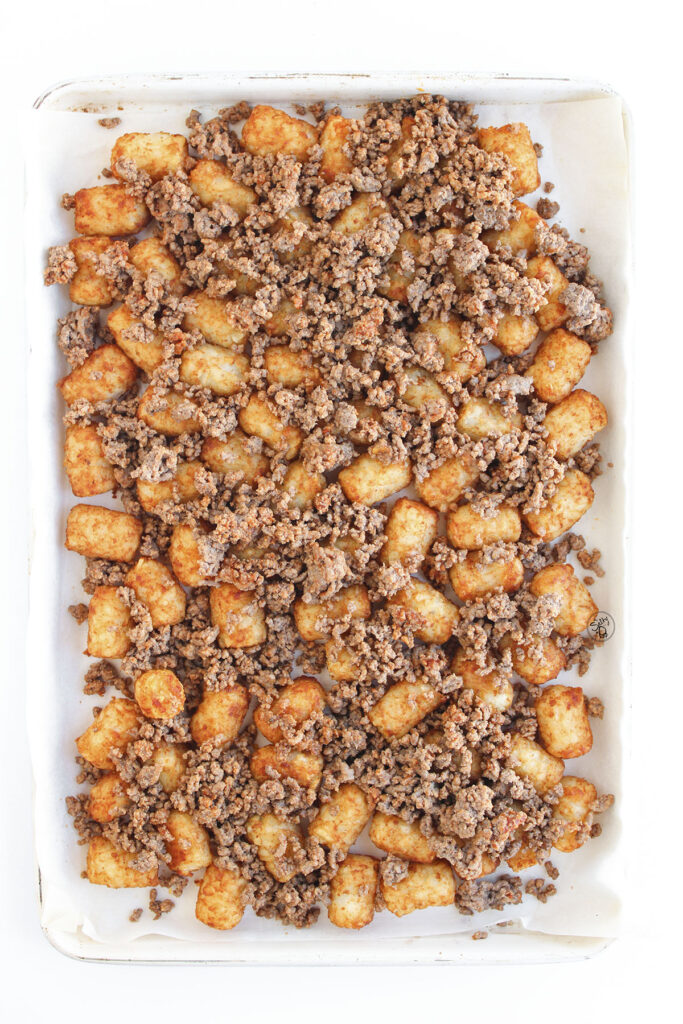 A sheet pan lined with parchment paper holding a layer of crispy golden tater tots topped with crumbled seasoned ground beef.