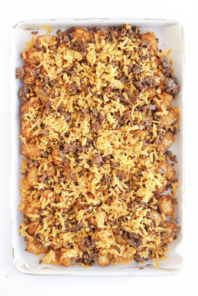 A sheet pan of tater tots and ground beef covered in a generous layer of shredded orange cheddar cheese before melting.
