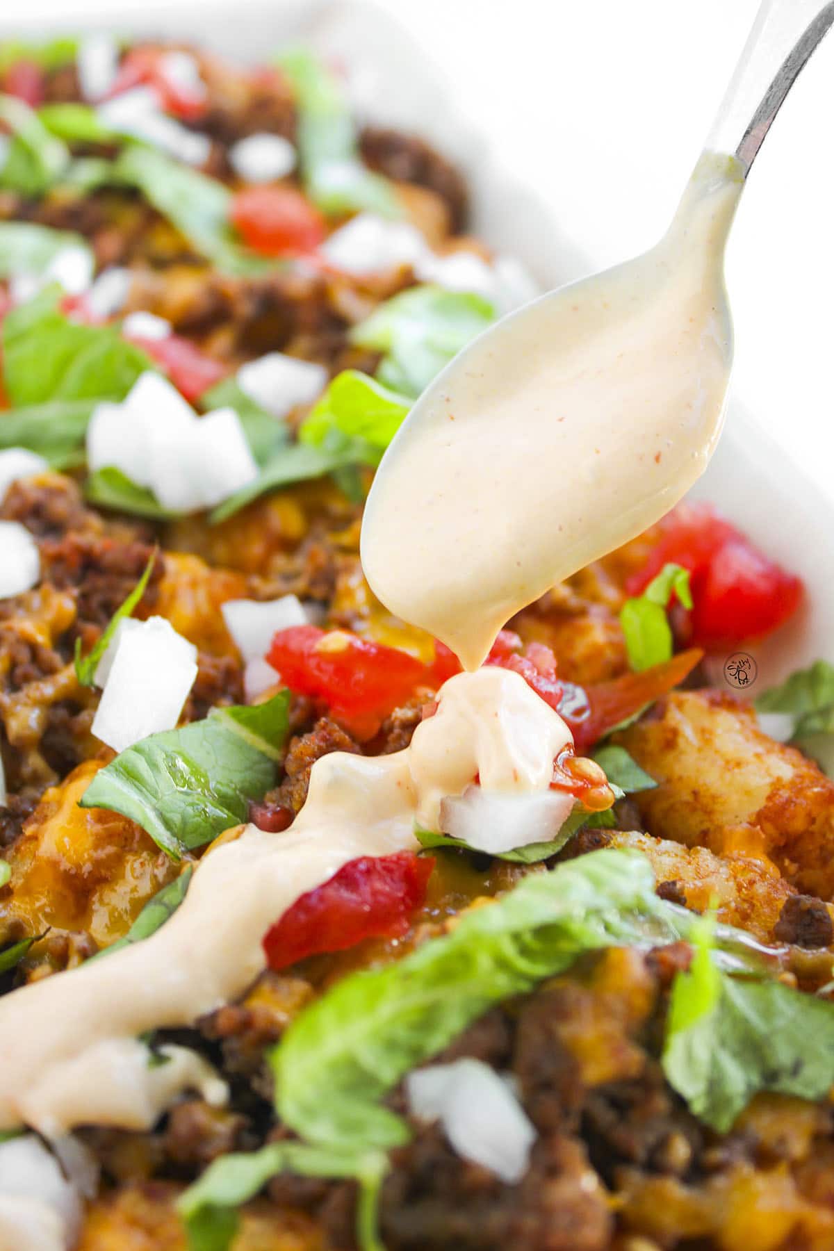 A close-up shot of a spoon drizzling creamy, orange-tinted burger sauce over a bed of loaded tater tots topped with beef, fresh vegetables, and pickles.