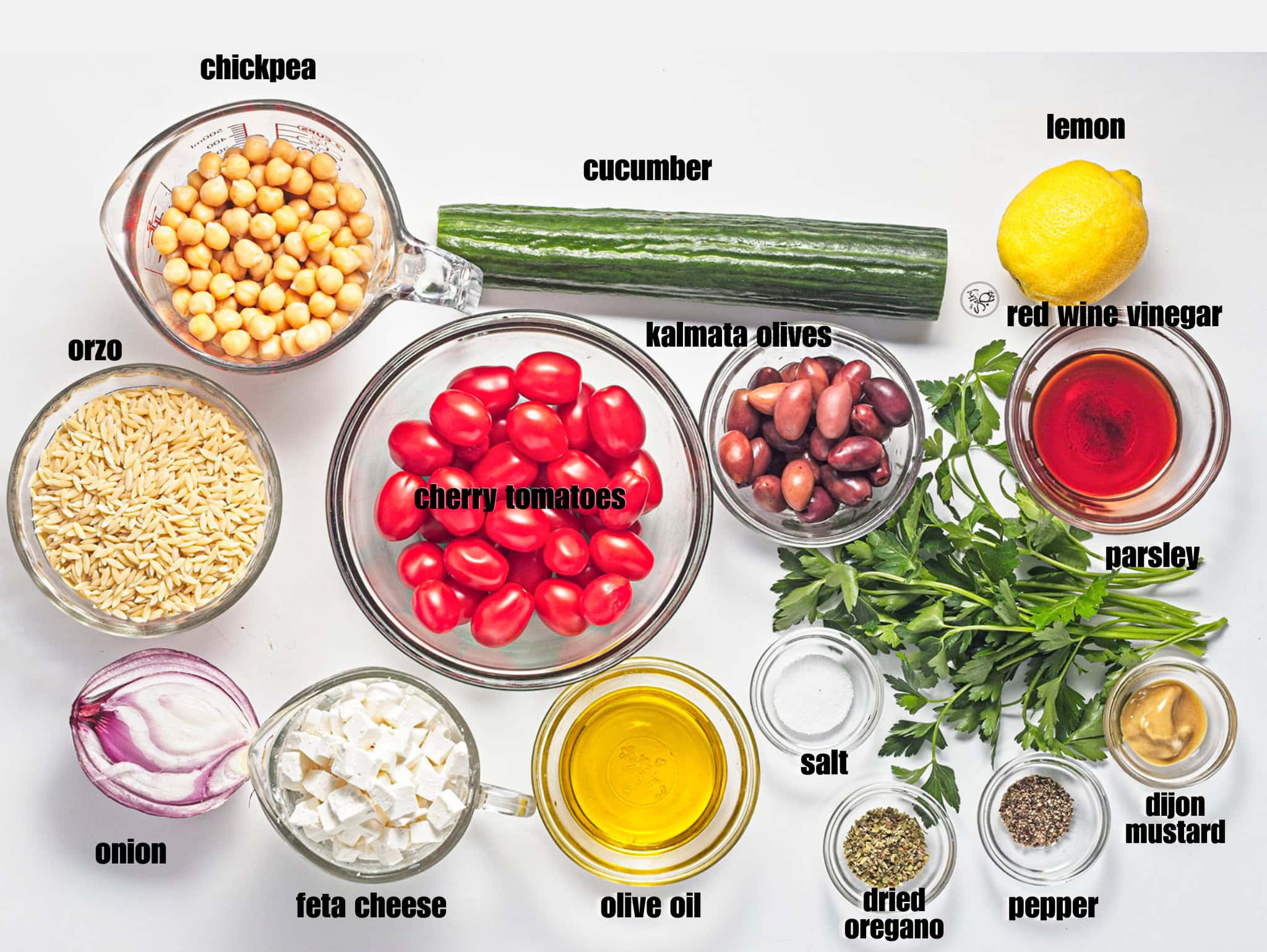 A top-down flat lay of raw ingredients for an orzo salad arranged on a white surface. The ingredients include cooked orzo pasta, canned chickpeas, cherry tomatoes, a long cucumber, Kalamata olives, cubed feta cheese, a red onion half, fresh parsley, a lemon, olive oil, red wine vinegar, dijon mustard, salt, pepper, and dried oregano.