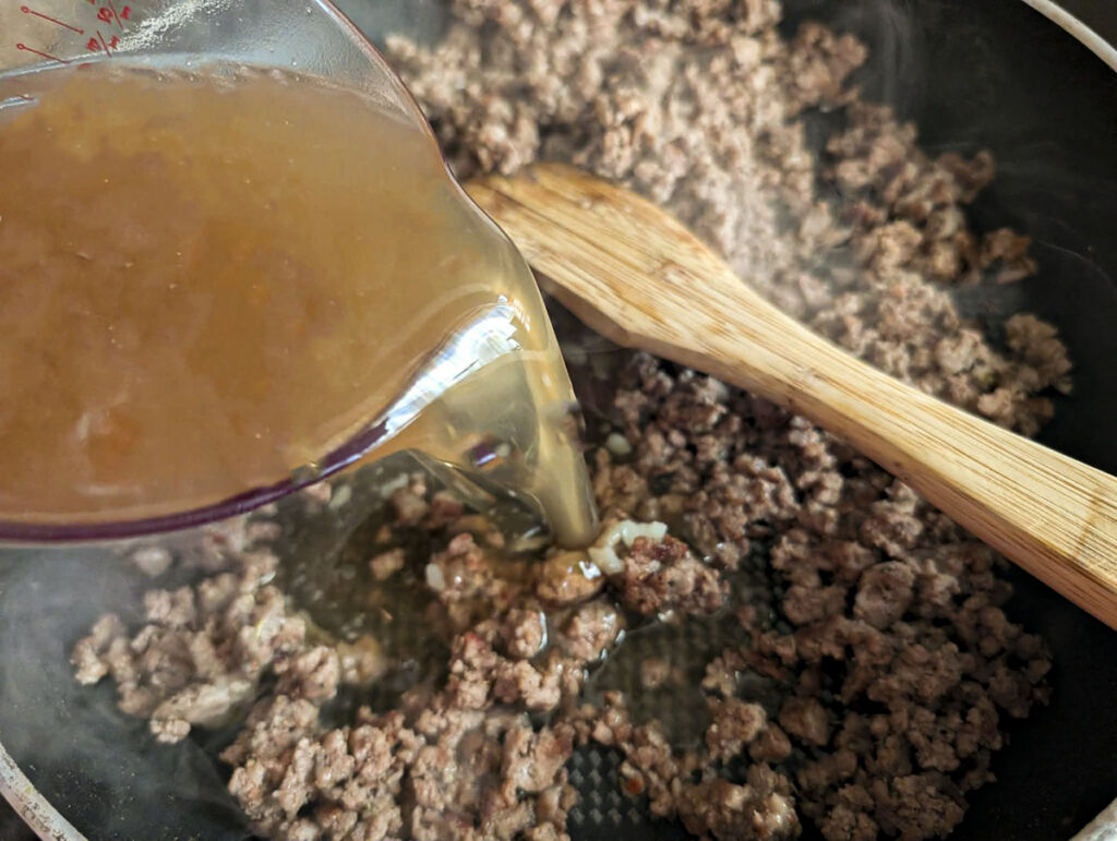 Pouring of beef broth from a glass measuring cup into a skillet of browned ground beef and flour.