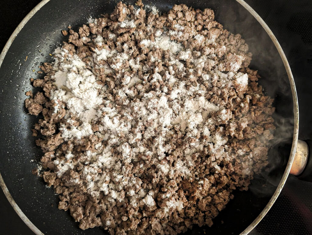 Ground beef browning in a black skillet with white all-purpose flour sprinkled over the top to create the gravy base.
