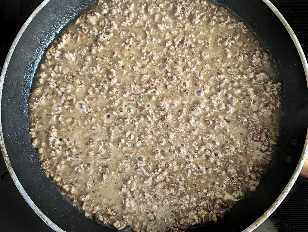 A close-up of hamburger gravy bubbling and simmering in a black pan as it thickens into a glossy sauce.