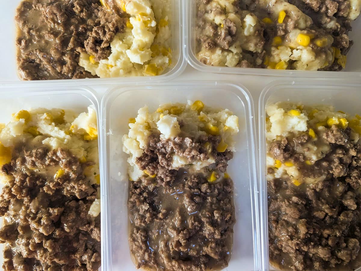 Five clear plastic meal-prep containers filled with individual servings of hamburger gravy over corn-mixed mashed potatoes, ready for the freezer.