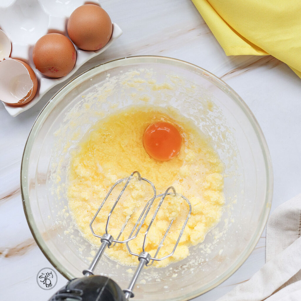 An overhead view of a clear glass bowl where a hand mixer is creaming together softened butter and sugar, with one egg cracked into the center.