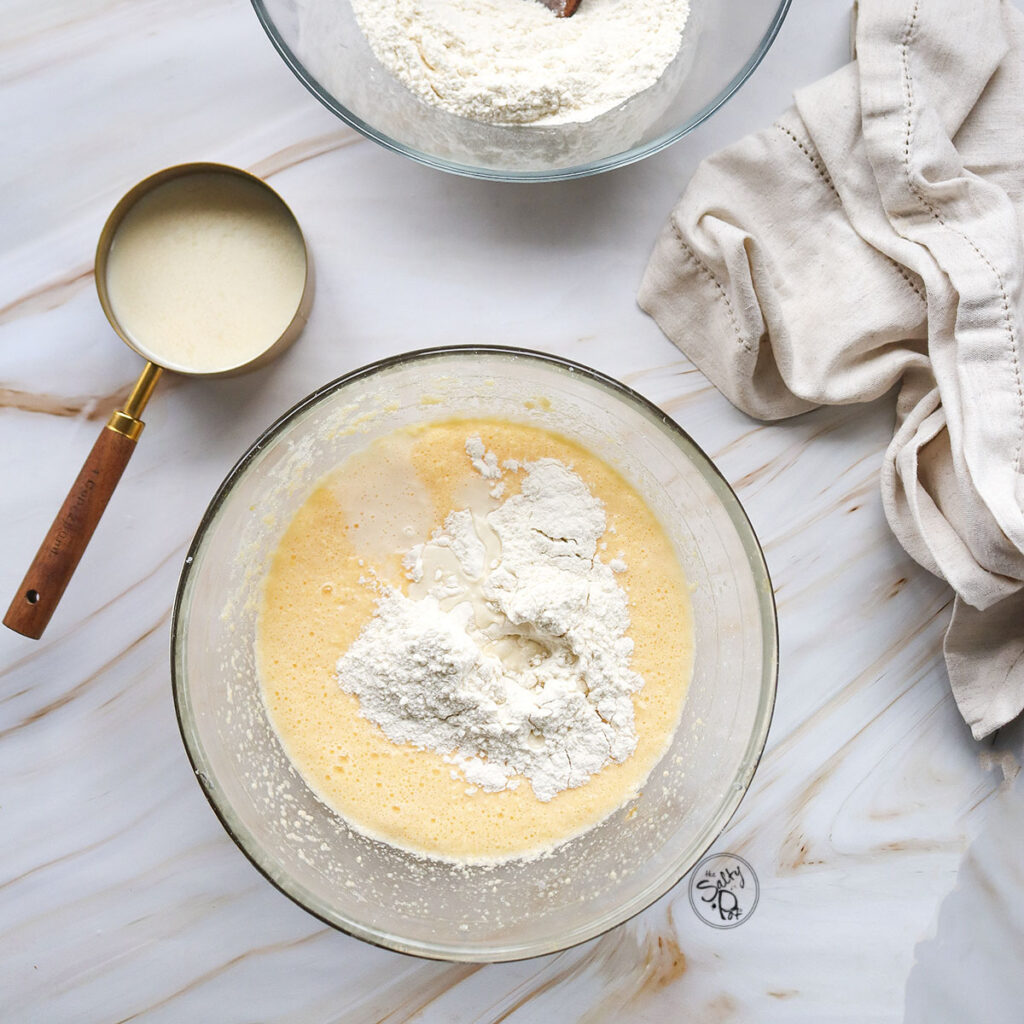 A glass bowl containing the wet cake batter with a fresh pile of white flour on top, placed next to a measuring cup of buttermilk and a beige linen towel.