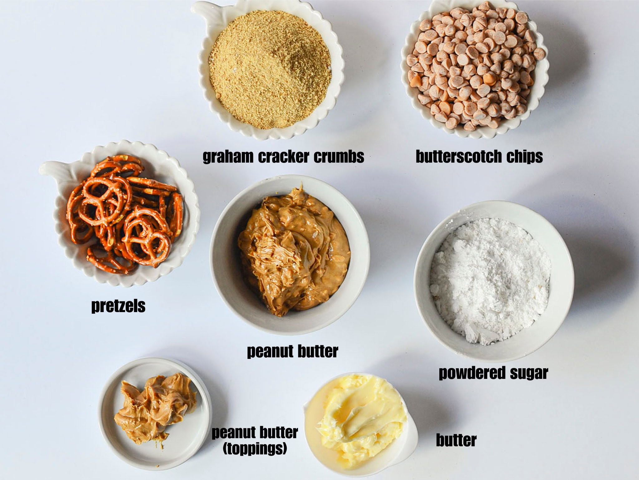A flat-lay view of ingredients in separate white bowls: graham cracker crumbs, pretzels, butterscotch chips, powdered sugar, butter, and peanut butter, all arranged on a white surface.