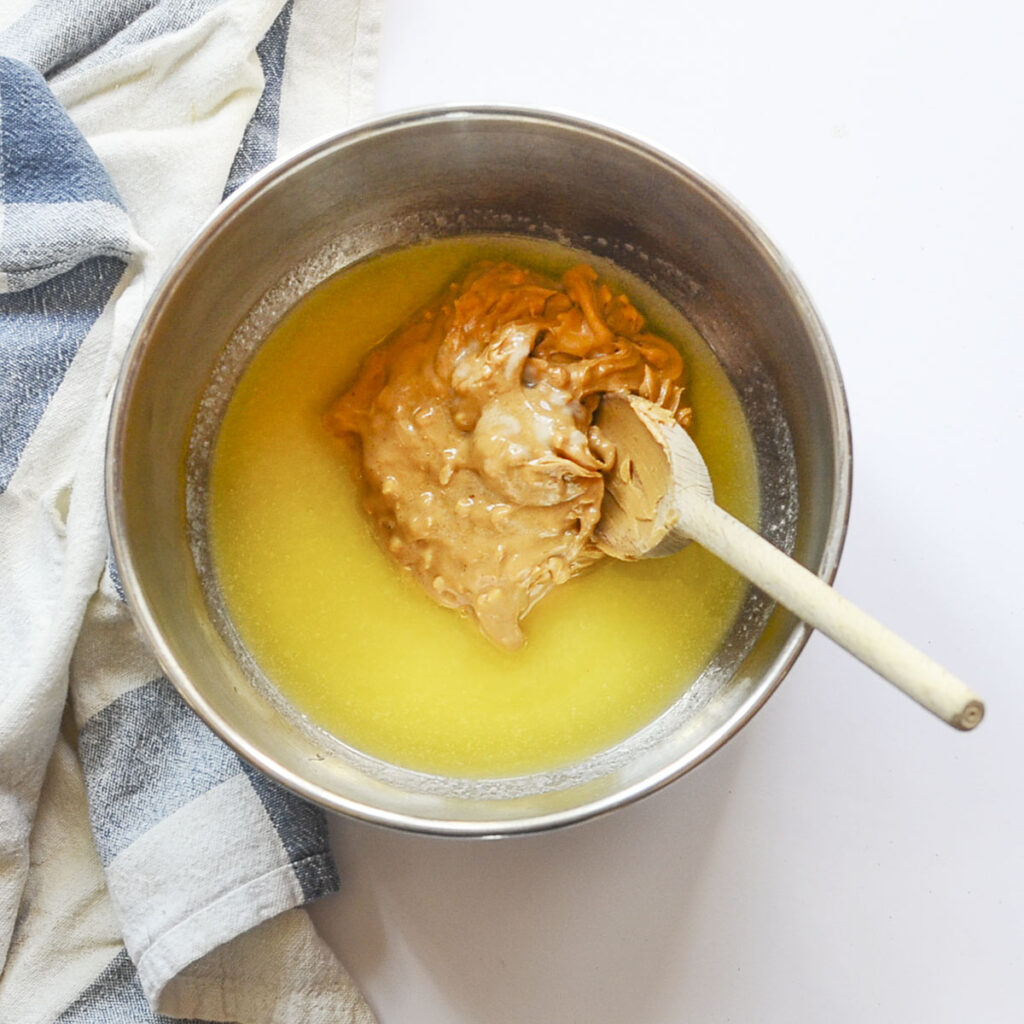 A stainless steel mixing bowl containing melted butter with a large scoop of peanut butter in the center, ready to be stirred with a wooden spoon.