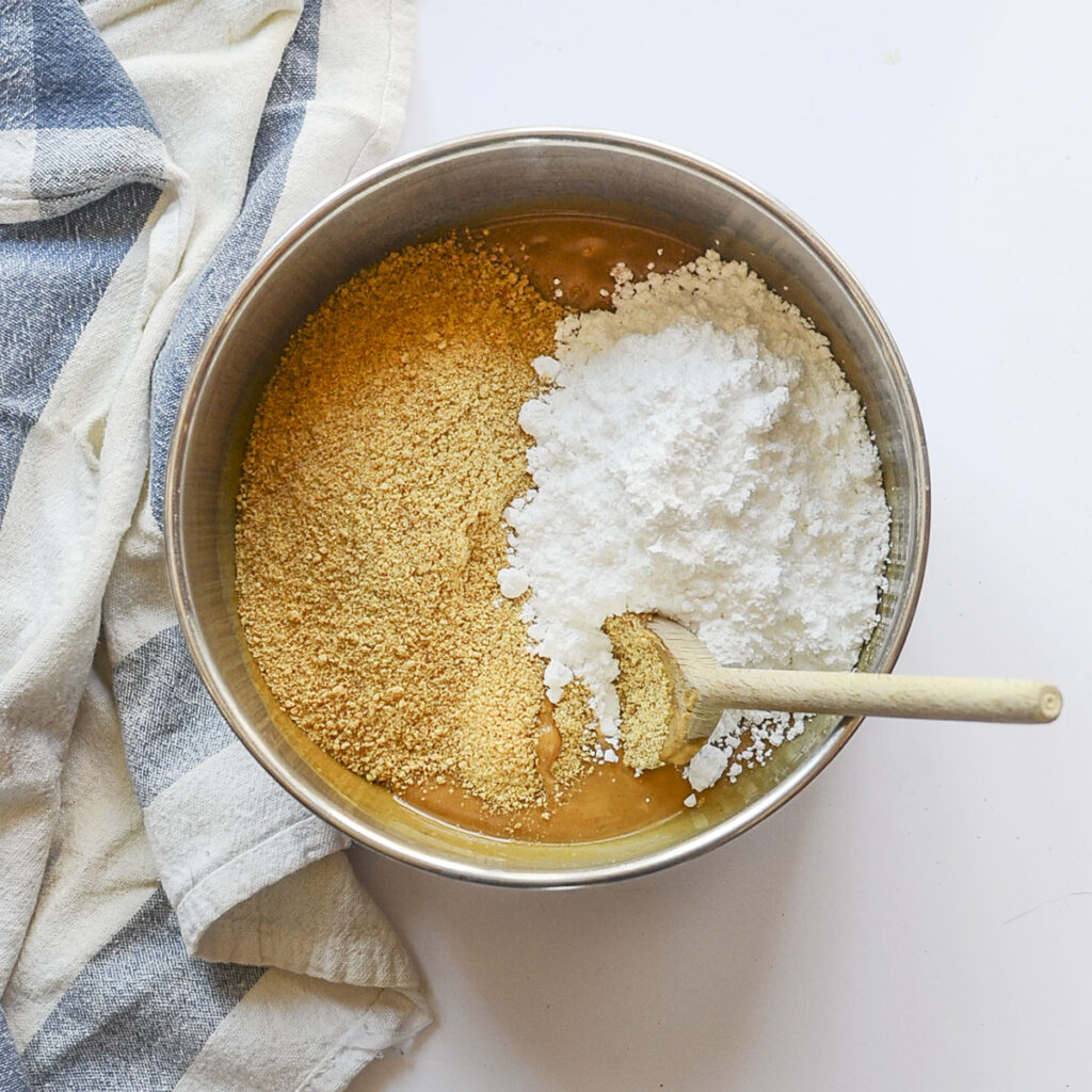 Powdered sugar and graham cracker crumbs being added to the peanut butter and butter mixture in the metal bowl.