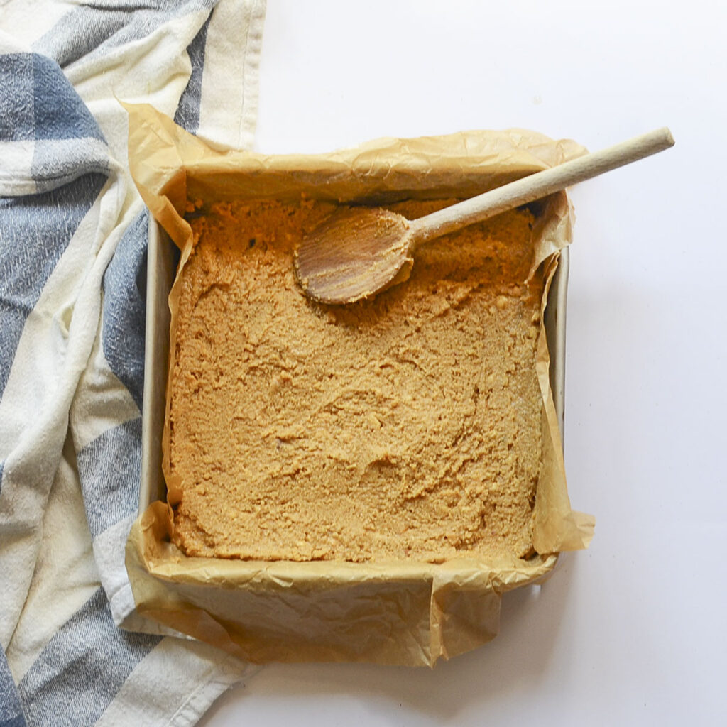 The thick peanut butter and graham cracker mixture pressed firmly into the bottom of a square baking pan lined with parchment paper.