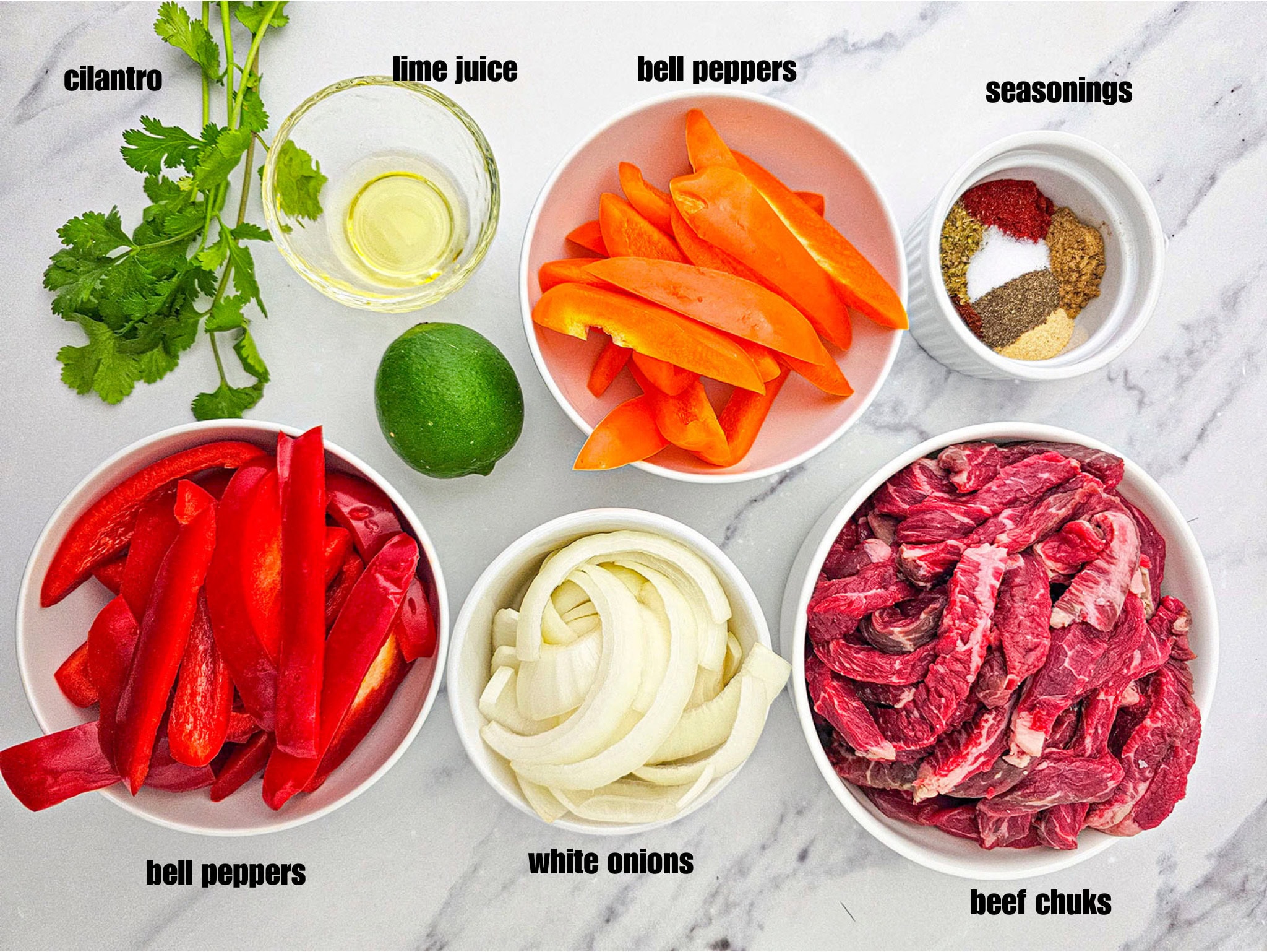 A top-down view of prepped ingredients in white bowls on a marble counter, including sliced beef roast, sliced red and orange bell peppers, sliced white onions, olive oil, a whole lime, fresh cilantro, and a small bowl of mixed spices.