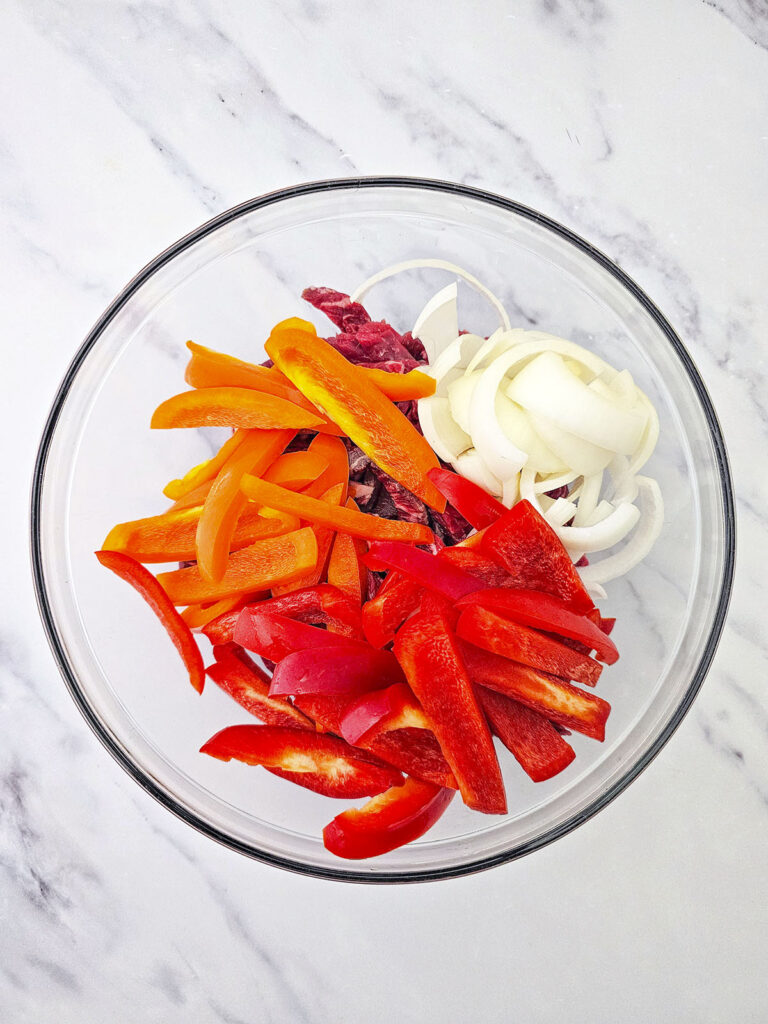 Sliced red and orange bell peppers and white onions layered over raw beef strips in a large clear glass mixing bowl.