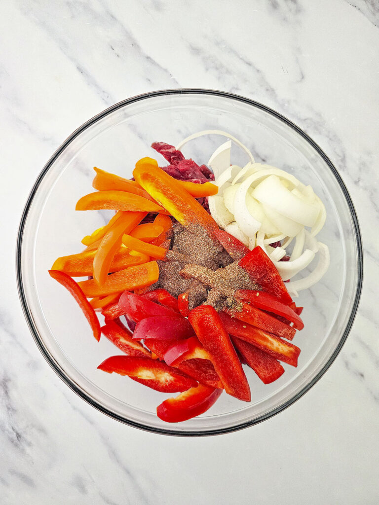 A heap of dark red and brown fajita seasoning blend sits on top of sliced peppers and onions in a glass bowl.