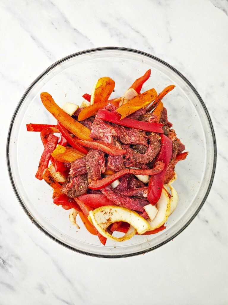 Sliced beef and colorful vegetables thoroughly coated in oil and spices, mixed together in a clear glass bowl.