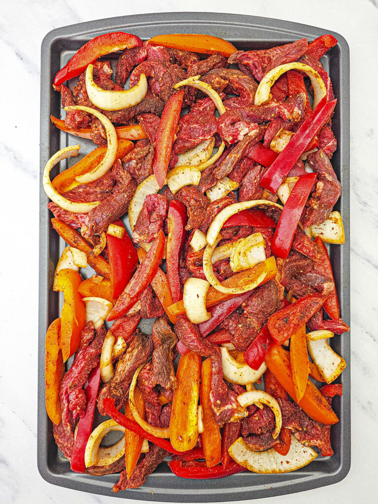 Raw, seasoned steak strips, peppers, and onions spread out in a single layer on a dark grey metal sheet pan, ready for the oven.
