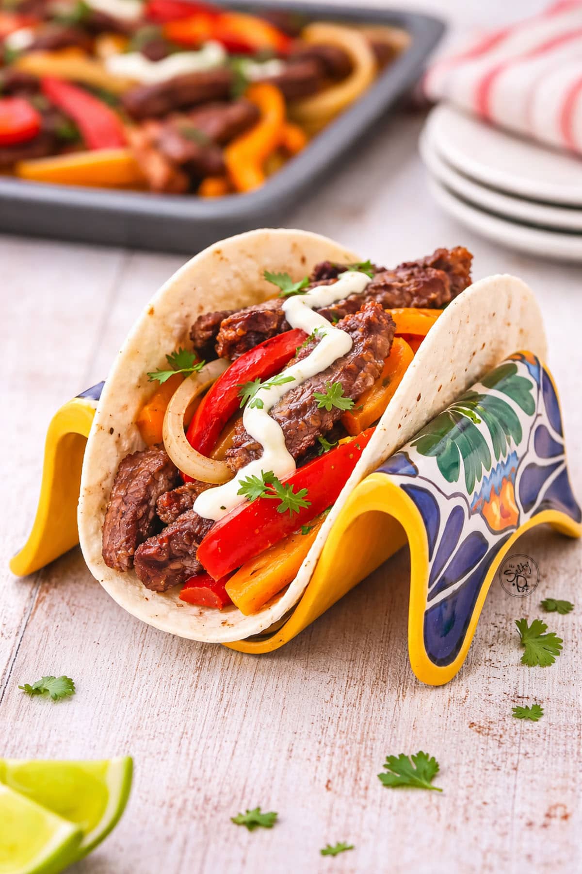 A close-up of a loaded steak fajita taco held in a colorful ceramic taco holder, topped with a drizzle of white cream and fresh cilantro, with the sheet pan visible in the background.