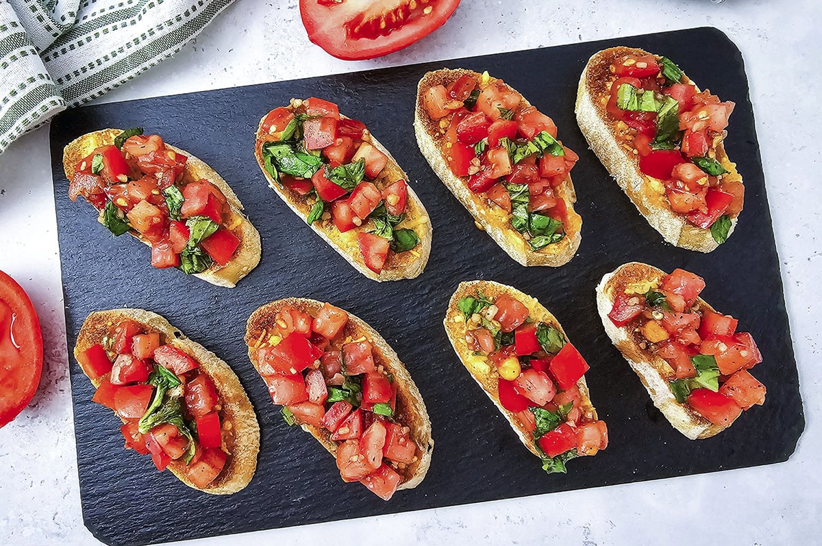 A row of assembled tomato bruschetta slices arranged on a long, dark slate serving platter.