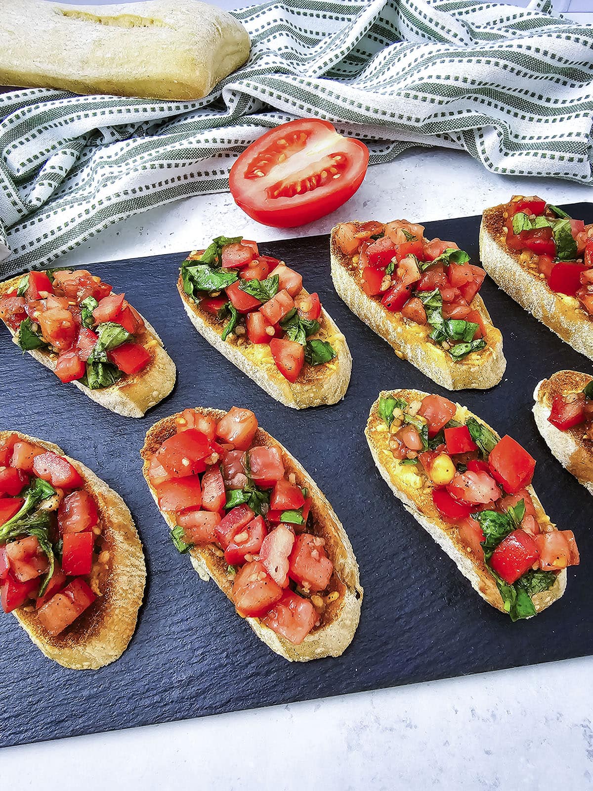 A row of assembled tomato bruschetta slices arranged on a dark slate serving platter.