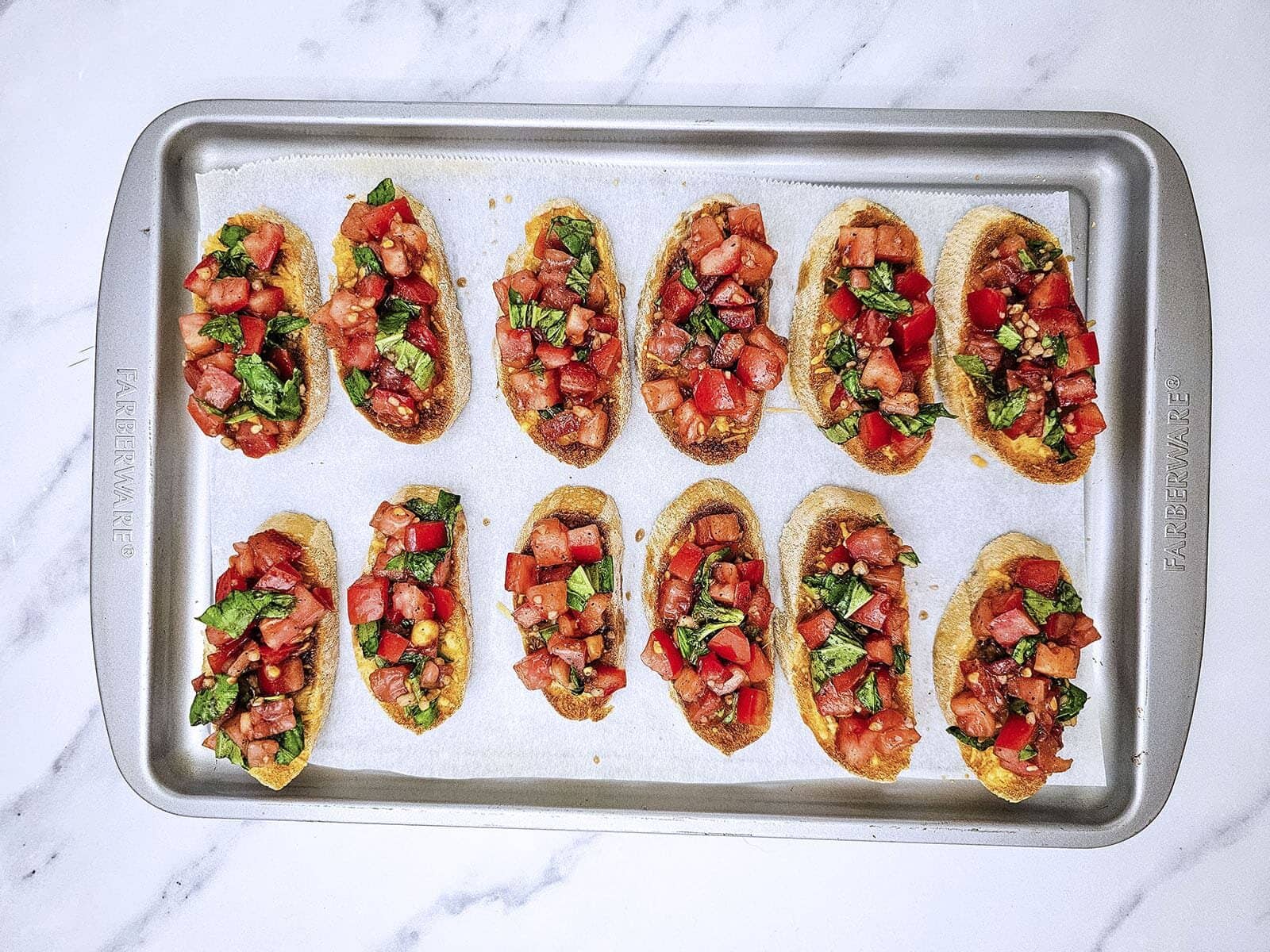 A row of toasted bread slices topped with the diced tomato mixture on a metal baking sheet.