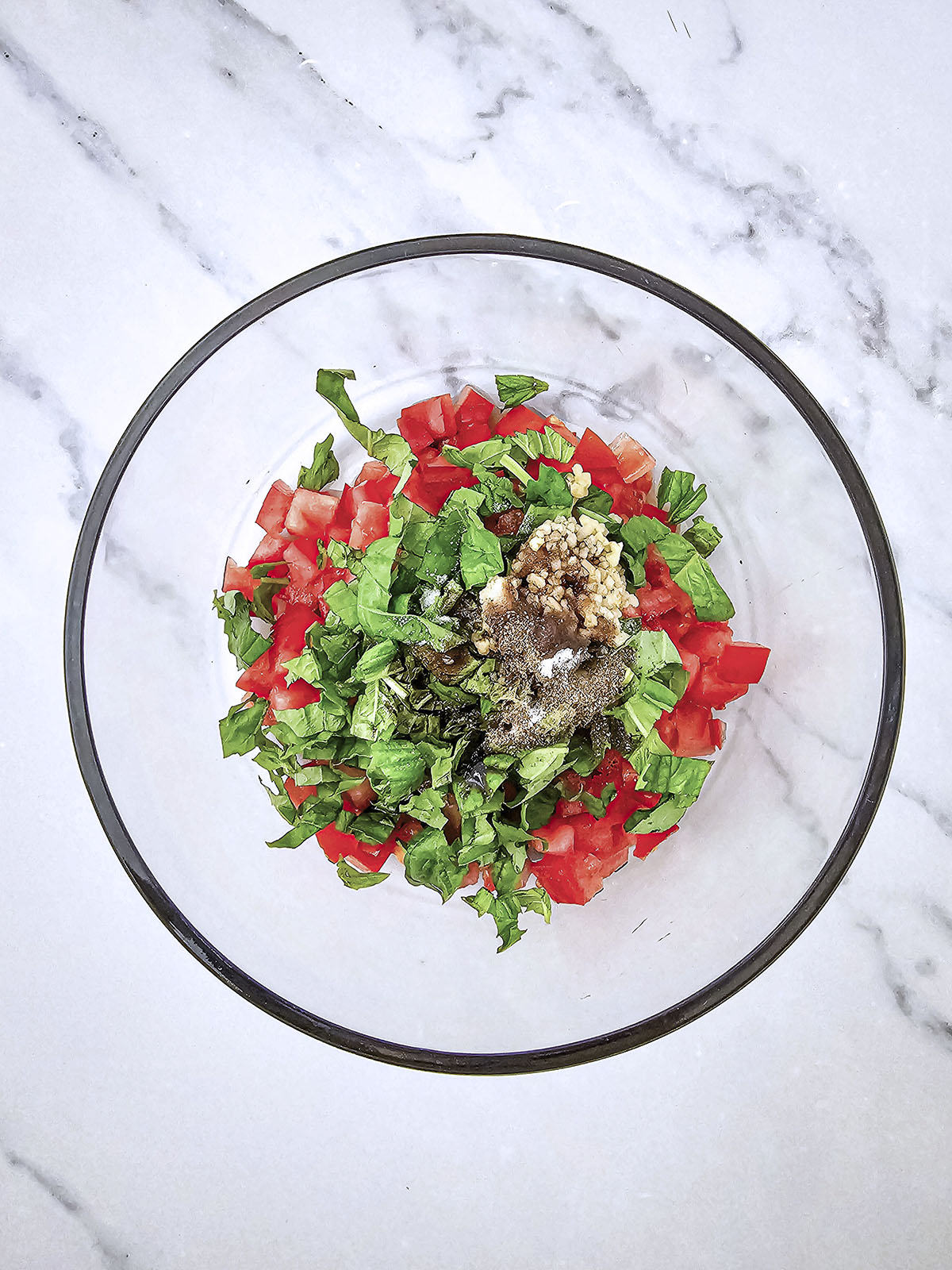 Unmixed diced tomatoes and chopped basil with the seasonings in a clear glass bowl.