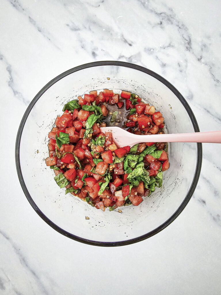 Diced tomatoes and chopped basils mixed with spices in a clear glass bowl.