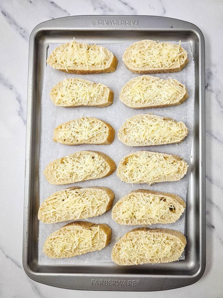 Slices of French bread topped with parmesan arranged on a metal baking sheet, ready to be toasted.