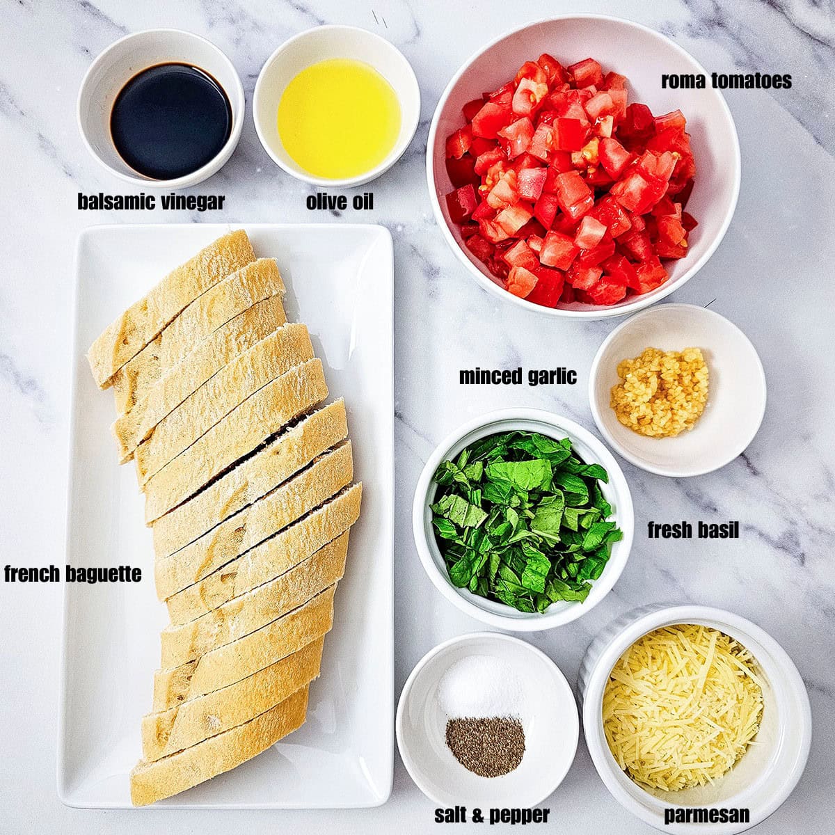 An overhead flat-lay of ingredients: a baguette, diced tomatoes, minced garlic, fresh basil, olive oil, and balsamic vinegar, parmesan cheese then salt and pepper