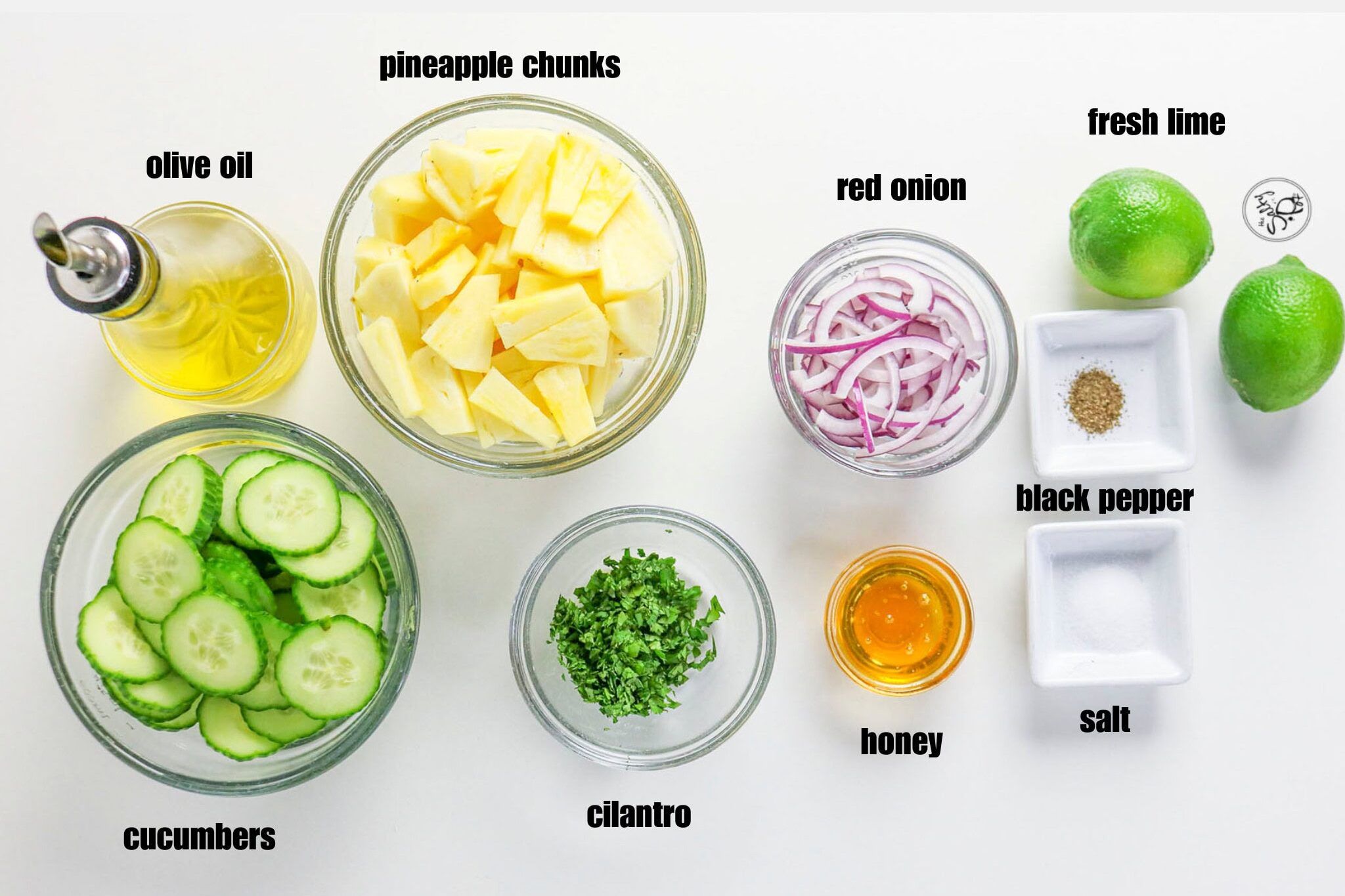 A flat-lay overview of prepped salad ingredients in separate small bowls on a white surface, including sliced cucumbers, pineapple chunks, sliced red onions, chopped cilantro, honey, oil, salt, pepper, and two limes.