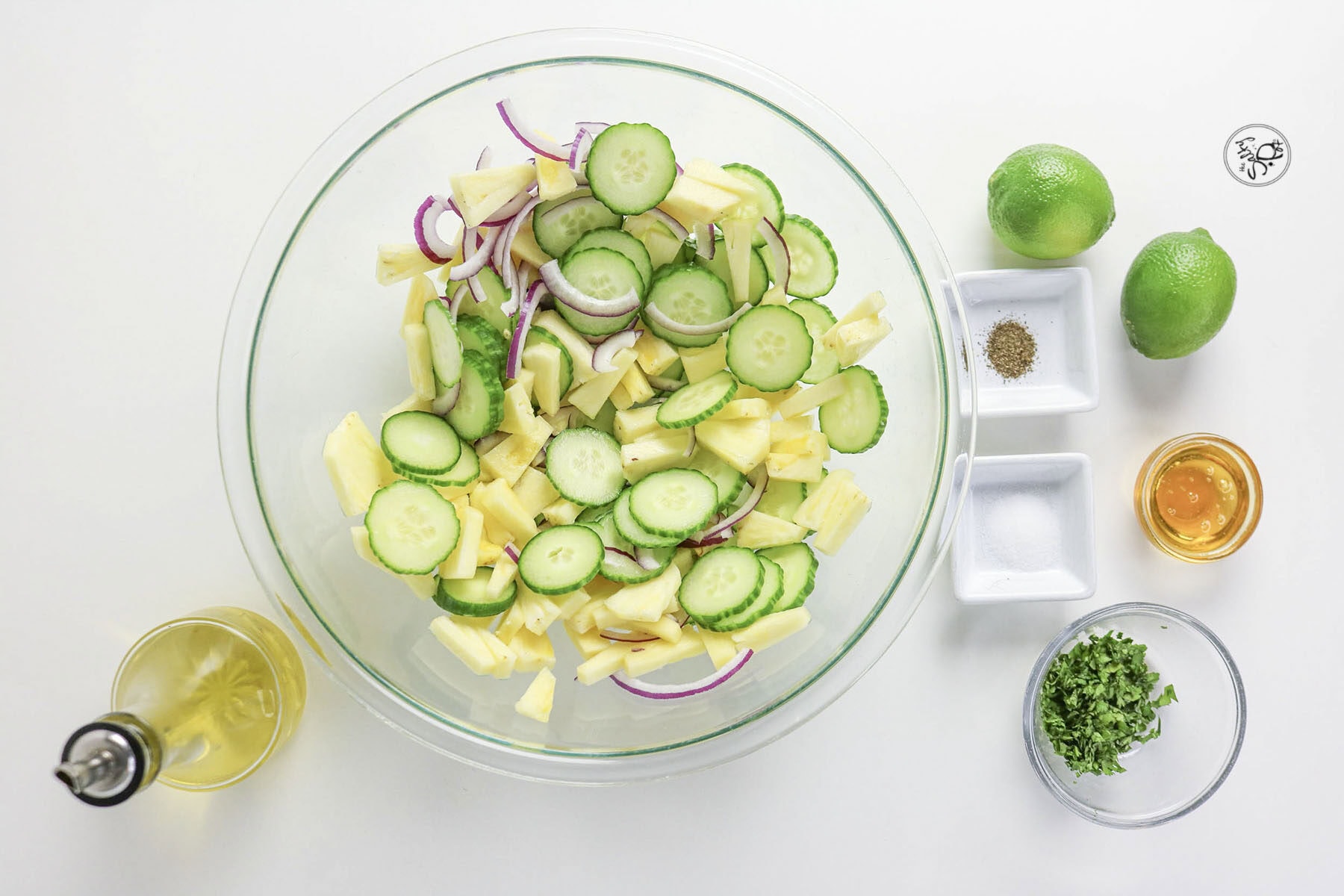 A top-down view of a glass bowl filled with chopped cucumber, pineapple, and red onion, surrounded by unmixed ingredients: chopped cilantro, oil, honey, small dishes of salt and pepper, and two whole limes.
