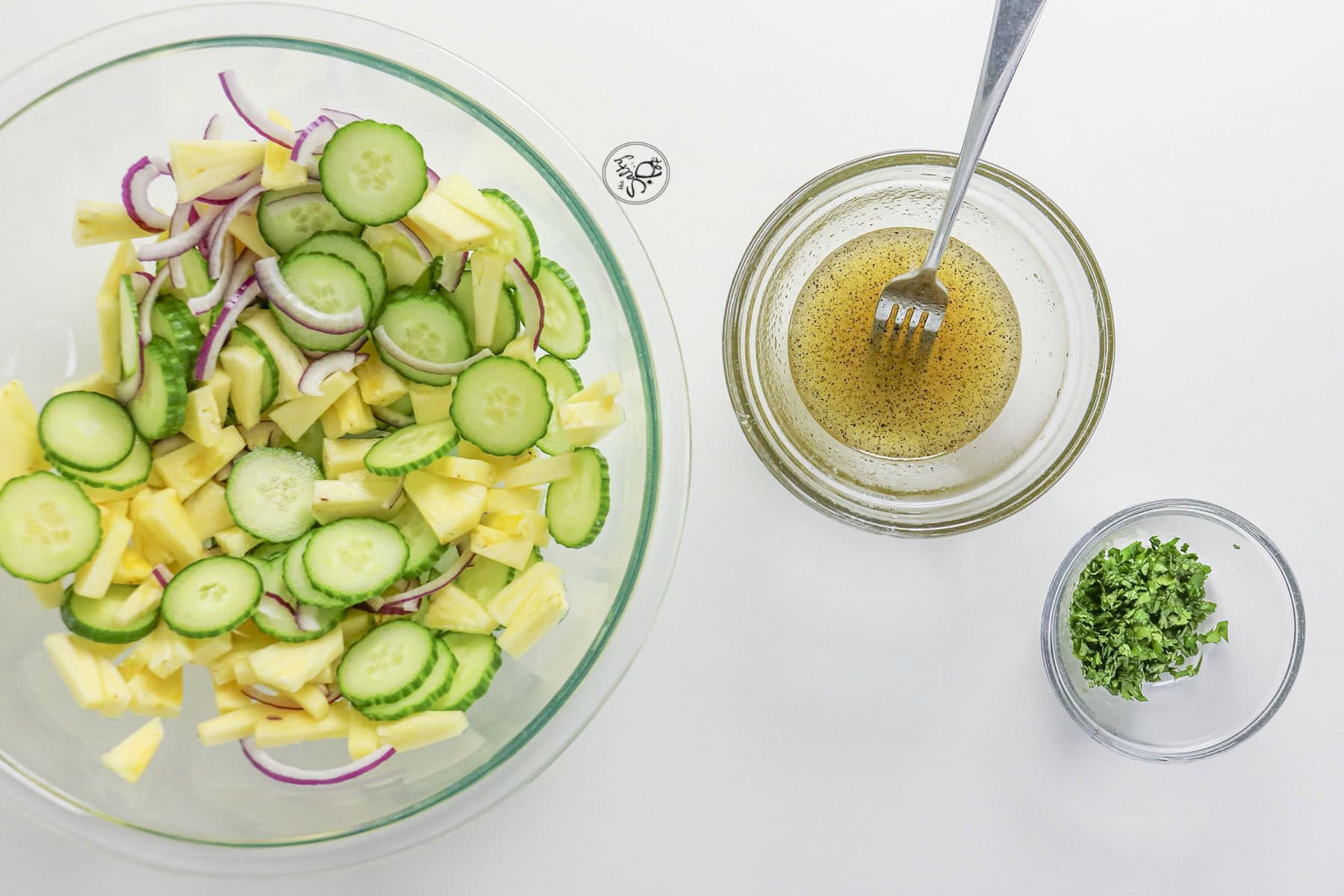 An overhead view showing a large glass bowl with cucumbers, pineapple, and red onion, alongside a smaller bowl of liquid dressing with a fork, and a tiny bowl of chopped cilantro.