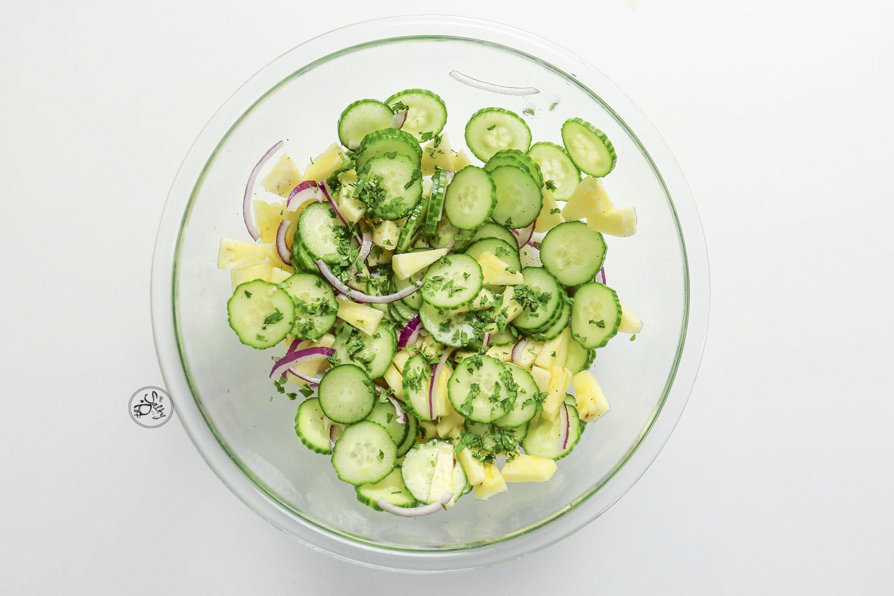 A top-down view of a clear glass bowl on a white background containing a mixed salad of sliced cucumbers, pineapple chunks, red onion, and chopped cilantro.