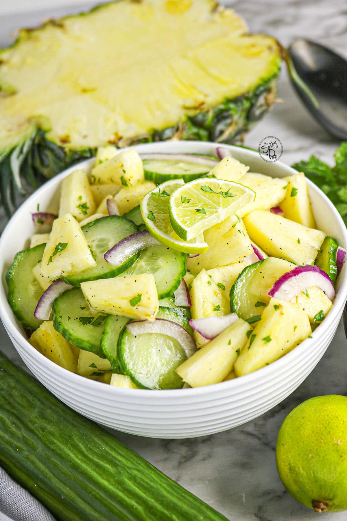 A close-up shot of a white bowl filled with a fresh salad of sliced cucumbers, pineapple chunks, and red onion, garnished with lime slices and chopped cilantro.
