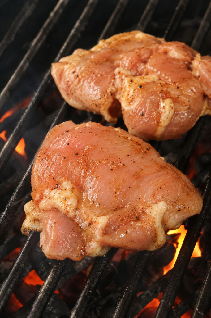 Seasoned boneless chicken thighs cooking on an outdoor grill with hot coals underneath.