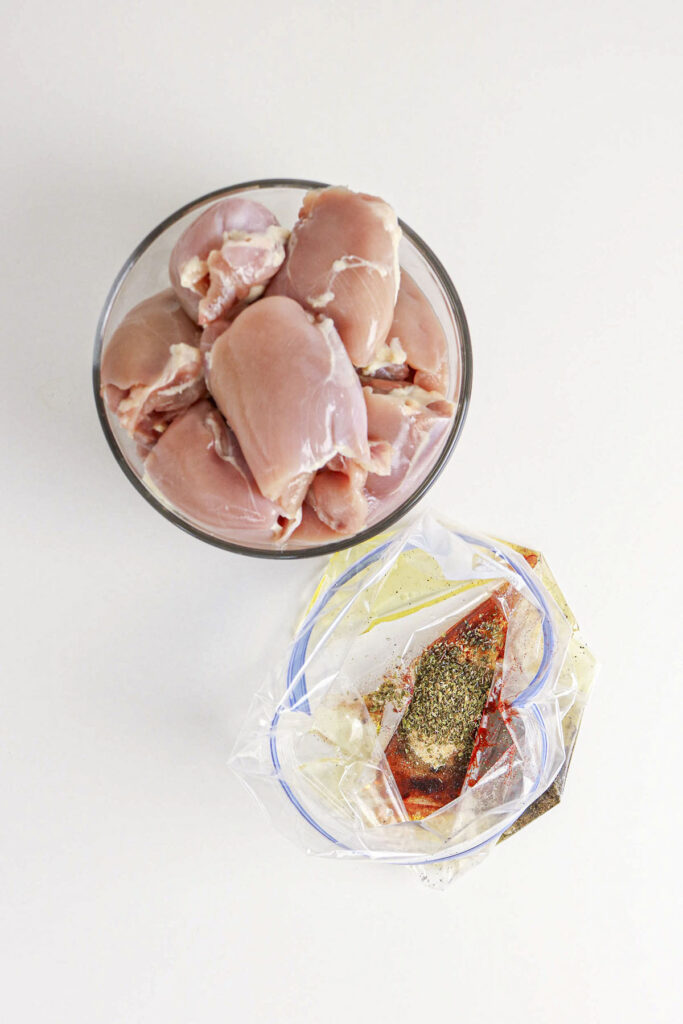 A bowl of raw chicken next to an open bag containing liquid and spices.