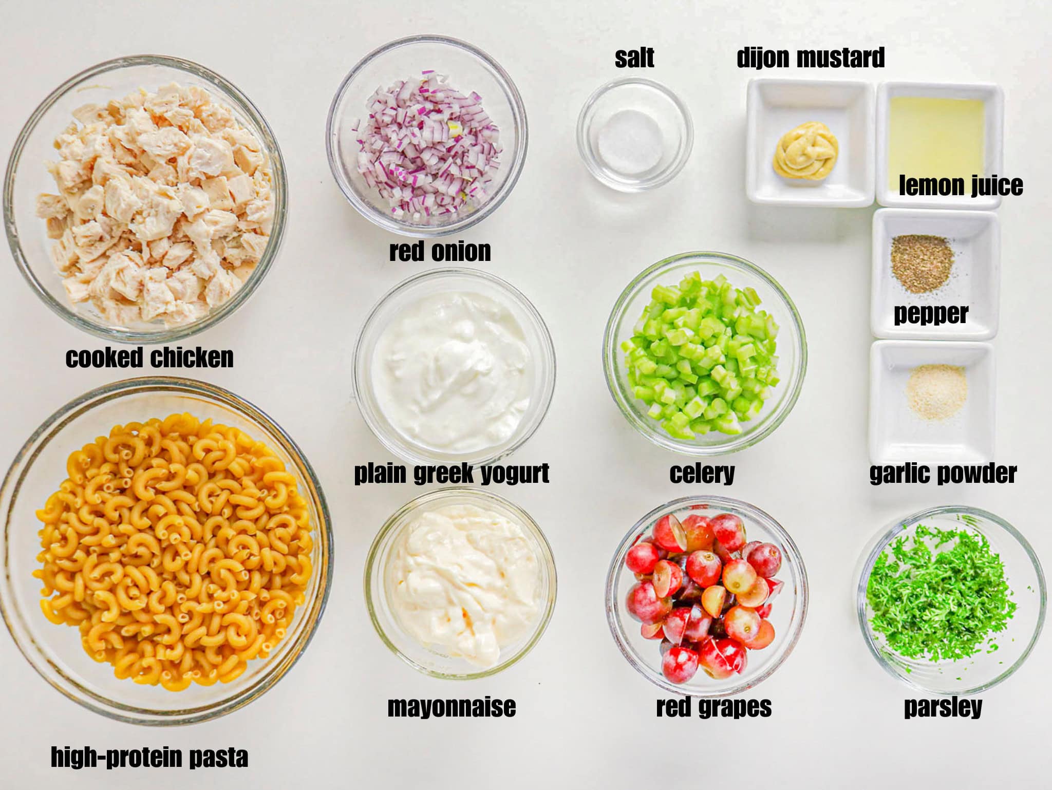 A top-down view of ingredients for a pasta salad arranged in separate bowls and small dishes on a white surface. The ingredients include cooked macaroni pasta, diced chicken, mayonnaise, Greek yogurt, diced red onion, halved red grapes, diced celery, chopped parsley, salt, garlic powder, black pepper, Dijon mustard, and lemon juice.