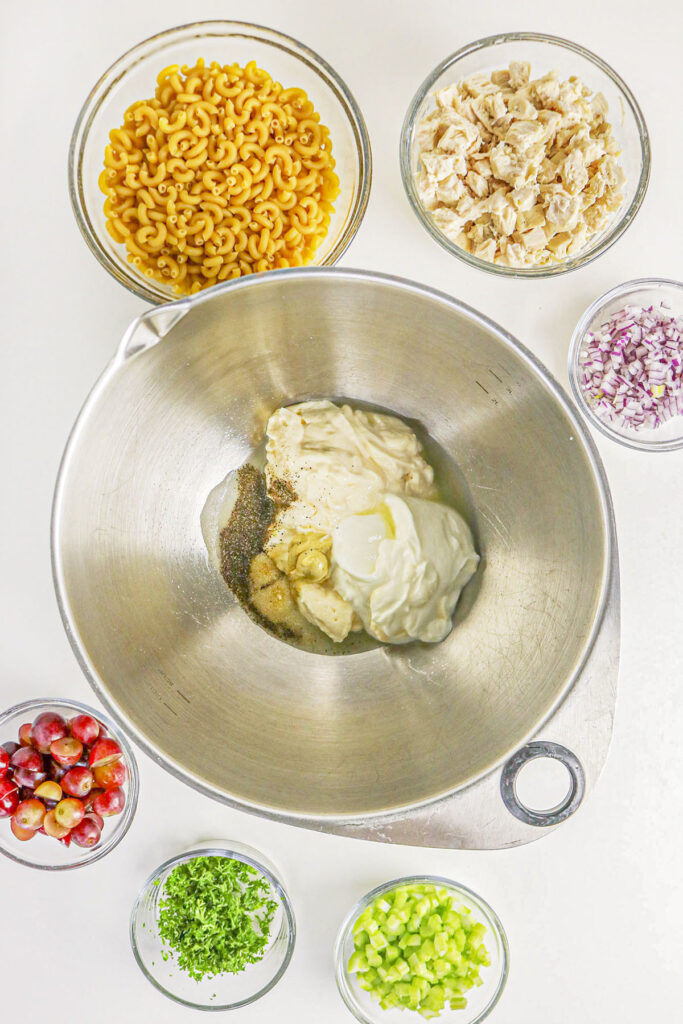 A top-down view of a large metal mixing bowl containing mayonnaise, Greek yogurt, mustard, lemon juice, and spices. Several smaller bowls containing pasta, diced chicken, grapes, celery, red onion, and parsley are arranged around the main mixing bowl on a white countertop.