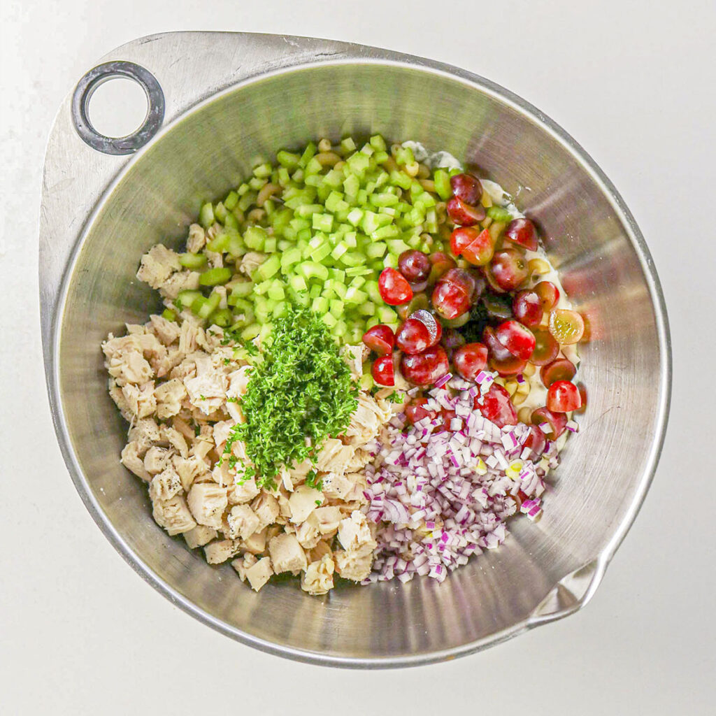 A top-down view of a large metal mixing bowl filled with ingredients before mixing. Diced chicken, chopped parsley, diced celery, halved red grapes, and diced red onion are arranged in distinct sections over a creamy dressing base.