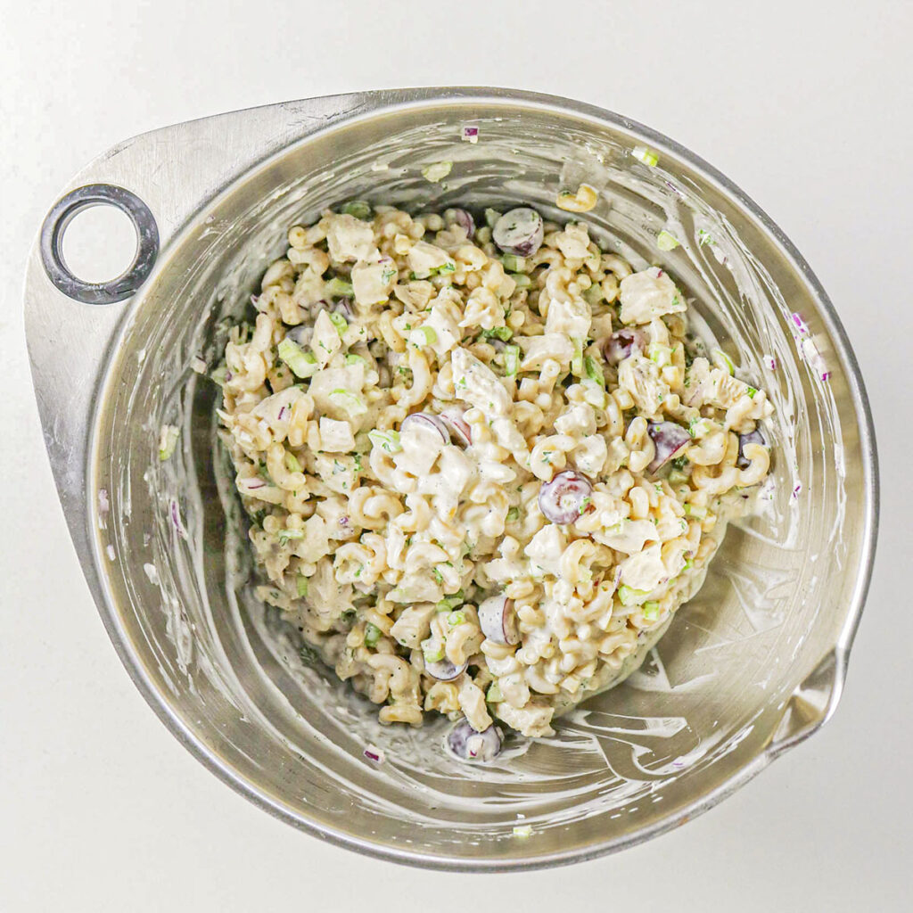 A top-down view of a large metal mixing bowl containing the fully mixed pasta salad, showing the macaroni, chicken, grapes, celery, and red onion coated uniformly in a white, creamy dressing.